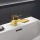 Waschtischarmatur in brushed Gold-Finish von Ideal Standard, mit flacher, breiter Auslaufkante, montiert auf einer weißen Oberfläche und schwarzer Kachelwand im Hintergrund.